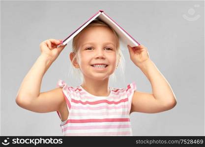 childhood and people concept - smiling little girl with roof of book on top of her head over grey background. little girl with roof of book on top of her head