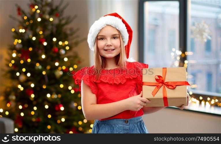 childhood and winter holidays concept - smiling girl in santa helper hat with gift box over christmas tree at home background. girl in santa hat with christmas gift at home