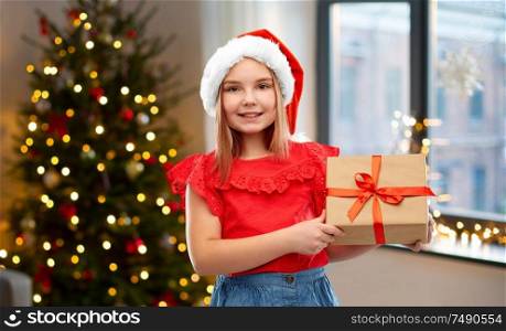 childhood and winter holidays concept - smiling girl in santa helper hat with gift box over christmas tree at home background. girl in santa hat with christmas gift at home