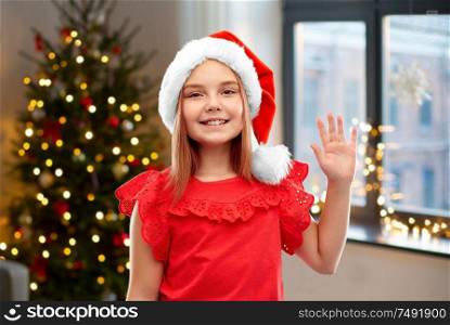 childhood and winter holidays concept - smiling girl posing in santa helper hat waving hand over christmas tree at home background. happy girl in santa helper hat waving hand at home