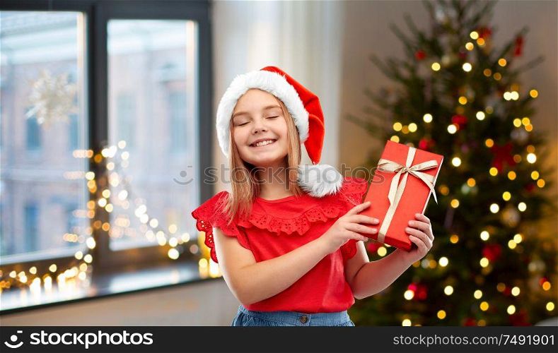 childhood and winter holidays concept - smiling happy girl in santa helper hat with gift box over christmas tree at home background. girl in santa hat with christmas gift at home