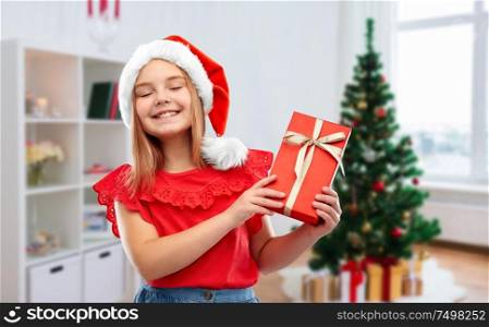 childhood and winter holidays concept - smiling happy girl in santa helper hat with gift box over christmas tree at home background. girl in santa hat with christmas gift at home
