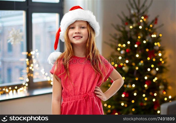 childhood and winter holidays concept - smiling red haired girl posing in santa helper hat over christmas tree at home background. happy red haired girl in santa helper hat at home