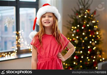 childhood and winter holidays concept - smiling red haired girl posing in santa helper hat over christmas tree at home background. happy red haired girl in santa helper hat at home