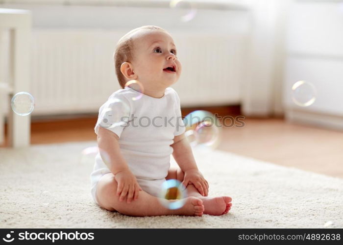 childhood, babyhood and people concept - happy little baby boy or girl sitting on floor with soap bubbles around at home