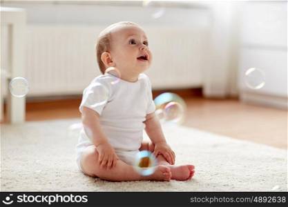 childhood, babyhood and people concept - happy little baby boy or girl sitting on floor with soap bubbles around at home