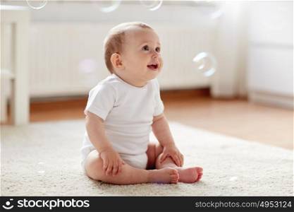 childhood, babyhood and people concept - happy little baby boy or girl sitting on floor with soap bubbles around at home. happy baby with soap bubbles at home