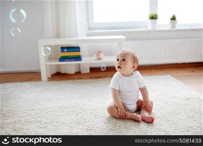 childhood, babyhood and people concept - happy little baby boy or girl sitting on floor with soap bubbles around at home. happy baby with soap bubbles at home