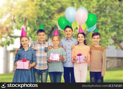 childhood, birthday, friendship and people concept - happy smiling children in party hats with gifts and balloons over garden background. happy children with gifts at birthday party