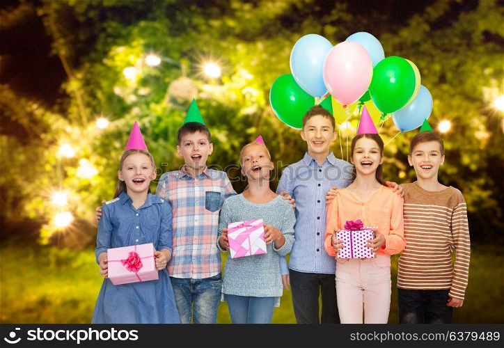 childhood, birthday, friendship and people concept - happy smiling children in party hats with gifts and balloons over festive lights at night garden background. happy children with gifts at birthday party