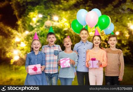 childhood, birthday, friendship and people concept - happy smiling children in party hats with gifts and balloons over festive lights at night garden background. happy children with gifts at birthday party