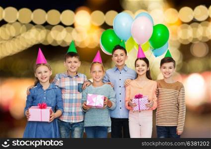 childhood, birthday, friendship and people concept - happy smiling children in party hats with gifts and balloons over festive lights background. happy children with gifts at birthday party