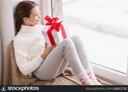 childhood, birthday, holidays and people concept - happy beautiful girl with christmas gift sitting on sill at home window