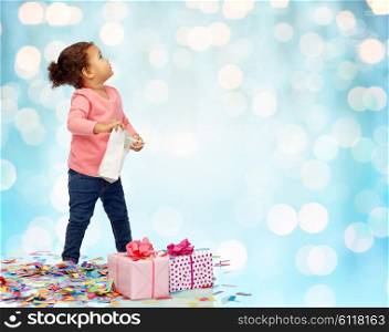 childhood, birthday, party, holidays and people concept - happy little african american baby girl with gift boxes and confetti playing with shopping bag looking up over blue holidays lights background