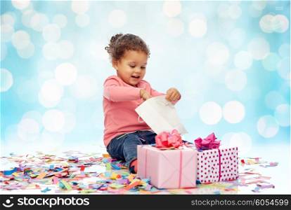 childhood, birthday, party, holidays and people concept - happy smiling little african american baby girl with gift boxes and confetti playing with shopping bag over blue lights background