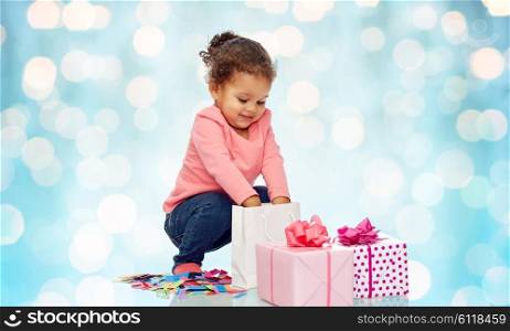 childhood, birthday, party, holidays and people concept - happy smiling little african american baby girl with gift boxes playing with confetti and shopping bag over blue holidays lights background
