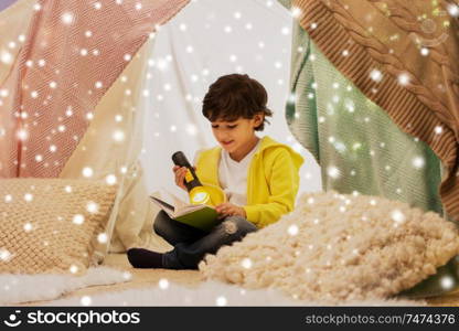 childhood, christmas and people concept - happy boy reading book with torch light in kids tent or teepee at home over snow. happy boy reading book in kids tent at home