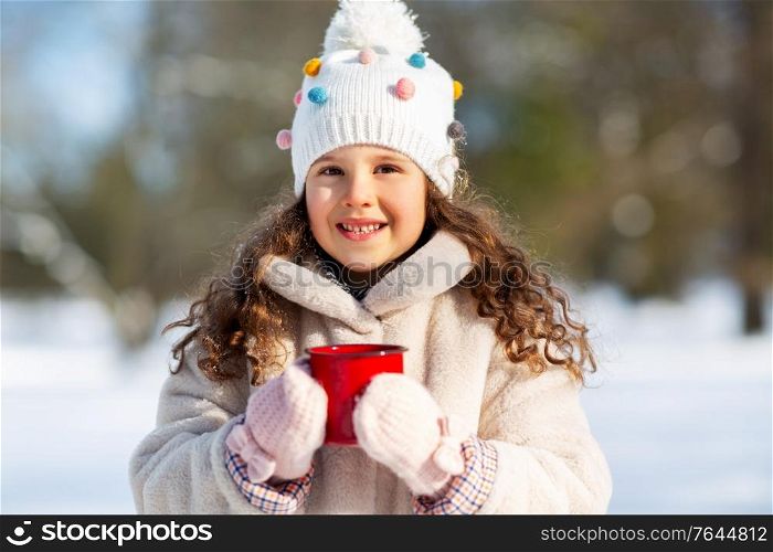 childhood, christmas and season concept - happy little girl with cup of hot tea in winter park. little girl with cup of hot tea in winter park