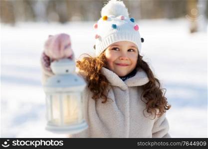 childhood, christmas and season concept - portrait of happy little girl in winter clothes with lantern outdoors at park. happy little girl with christmas lantern in winter