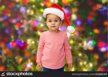 childhood, christmas, holidays and people concept - beautiful little african american baby girl in santa hat over lights background