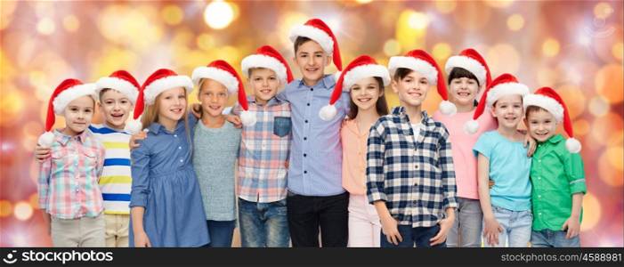 childhood, christmas, holidays and people concept - happy smiling children in santa hats hugging over lights background