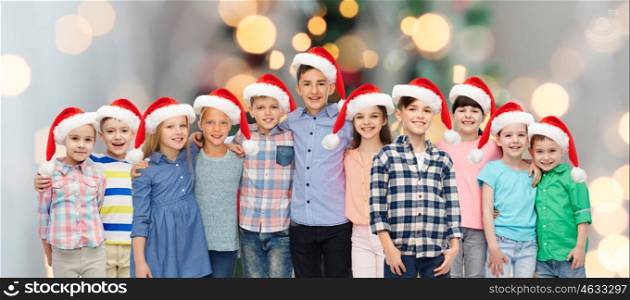 childhood, christmas, holidays and people concept - happy smiling children in santa hats hugging over lights background
