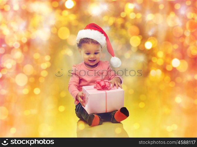 childhood, christmas, holidays and people concept - happy smiling little african american baby girl in santa hat with gift box sitting on floor over lights background