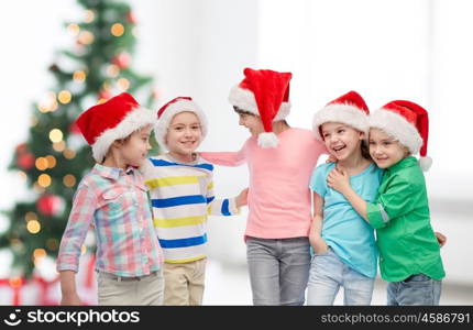 childhood, christmas, holidays, friendship and people concept - group of happy smiling little children in santa hats hugging over room with tree