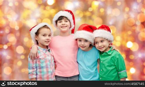childhood, christmas, holidays, friendship and people concept - group of happy smiling little children in santa hats hugging over holidays lights background