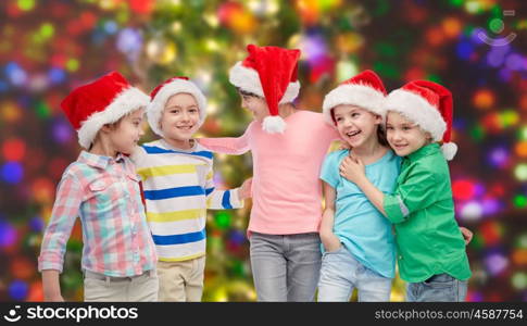 childhood, christmas, holidays, friendship and people concept - group of happy smiling little children in santa hats hugging over holidays lights background