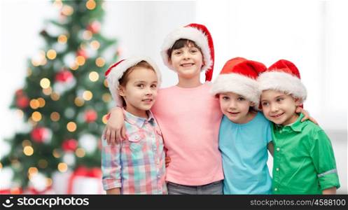 childhood, christmas, holidays, friendship and people concept - group of happy smiling little children in santa hats hugging over room with tree