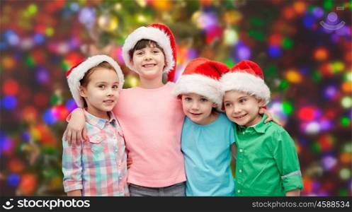 childhood, christmas, holidays, friendship and people concept - group of happy smiling little children in santa hats hugging over holidays lights background
