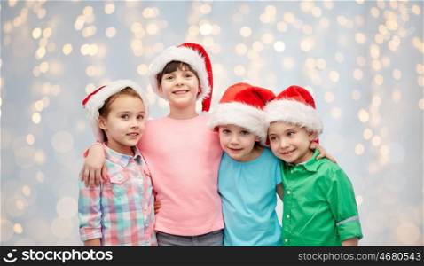 childhood, christmas, holidays, friendship and people concept - group of happy smiling little children in santa hats hugging over lights background