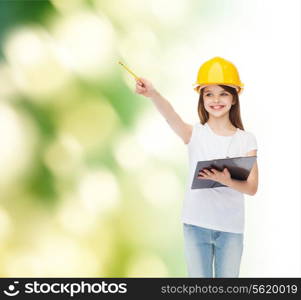 childhood, construction, architecture, building and people concept - smiling little girl in protective helmet with clipboard pointing pencil