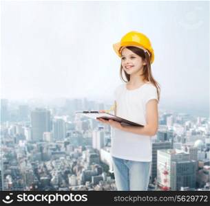 childhood, construction, architecture, building and people concept - smiling little girl in protective helmet with clipboard making notes