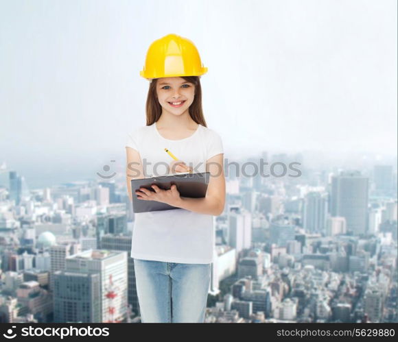 childhood, construction, architecture, building and people concept - smiling little girl in protective helmet with clipboard making notes