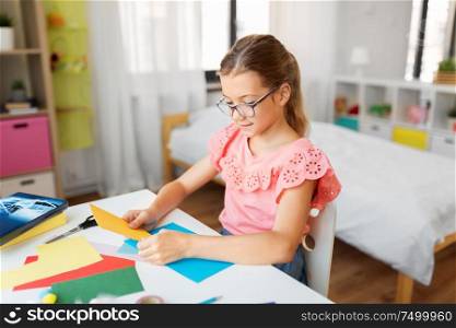 childhood, creativity and hobby concept - creative girl in glasses with color paper sitting at table at home. girl with color paper sitting at table at home