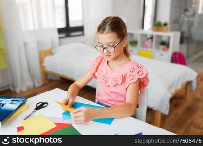 childhood, creativity and hobby concept - creative girl in glasses with color paper sitting at table at home. girl with color paper sitting at table at home