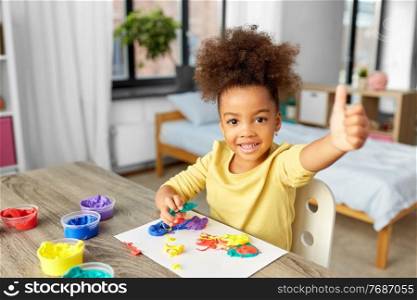 childhood, creativity and hobby concept - little african american girl playing with modeling clay and showing thumbs up at home. little girl with modeling clay playing at home