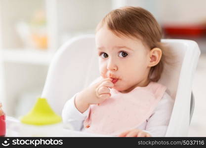 childhood, eating, feeding and people concept - happy little baby girl with bib sitting in highchair at home. happy baby girl sitting in highchair at home