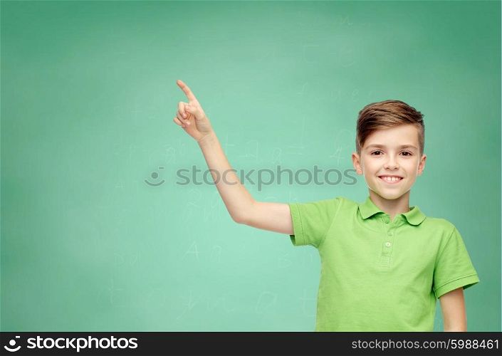 childhood, education and people concept - happy smiling boy in green polo t-shirt pointing finger up over green school chalk board background