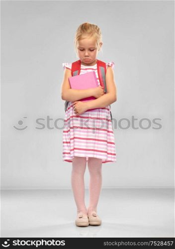 childhood, education and people concept - sad little student girl with school bag and book over grey background. sad little student girl with school bag and book