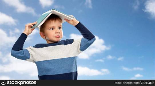 childhood, education and reading concept - little boy with roof of book on top of his head over blue sky and clouds background. little boy with roof of book on his head over sky