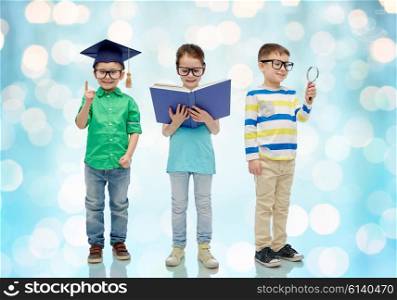 childhood, education, knowledge and people concept - happy little children in eyeglasses with book, magnifying glass and mortar board over blue holidays lights background