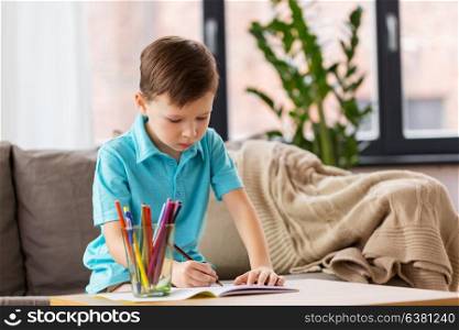 childhood, education, leisure and people concept - happy little boy with notebook and pencils drawing at home. boy with notebook and pencils drawing at home