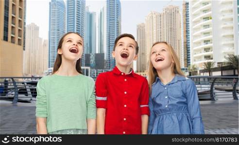 childhood, emotions, travel, tourism and people concept - happy amazed boy and girls looking up with open mouths over dubai city street background