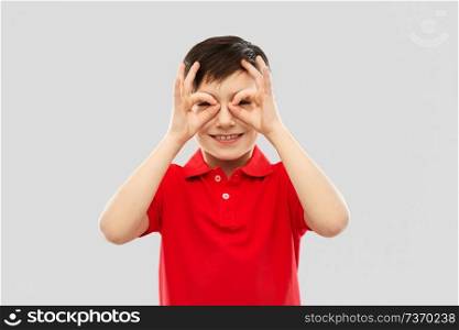 childhood, expressions and fun concept - happy little boy in red polo t-shirt looking through finger glasses over grey background. boy in red t-shirt looking through finger glasses