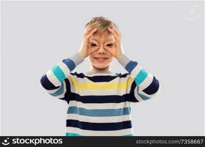 childhood, expressions and people concept - little boy in striped pullover looking through finger glasses over grey background. boy in pullover looking through finger glasses