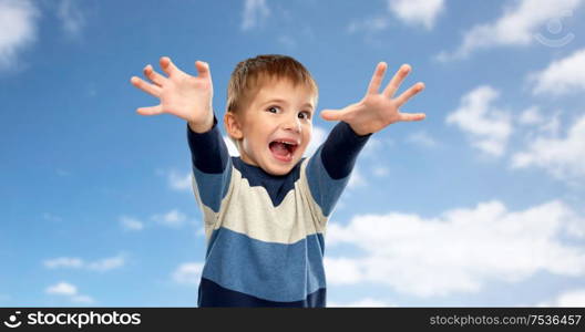 childhood, expressions and people concept - little boy in striped pullover making scary faces over blue sky and clouds background. little boy making scary faces over sky background