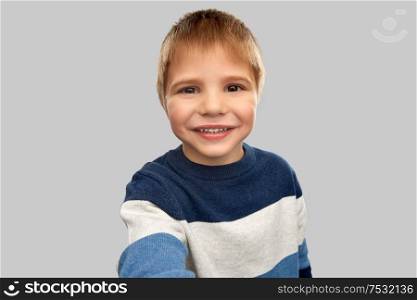 childhood, expressions and people concept - portrait of nice little boy in striped pullover taking selfie over grey background. happy little boy in striped pullover taking selfie